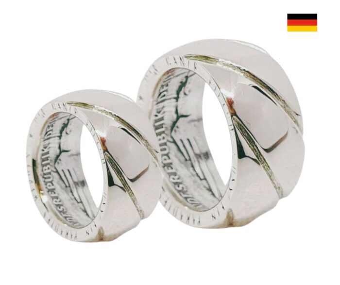 Bereit für die ewige Liebe: Entdecken Sie unsere handgefertigten Partner- und Eheringe aus originalen 10 DM Silbermünzen. Jeder Ring ist ein Unikat und wird mit einer individuellen Gravur versehen, um Ihre ganz persönliche Geschichte zu erzählen. Details: Gravurtext innen: Verleihen Sie Ihrem Ring eine persönliche Note mit einem individuellen Gravurtext. Material: 925 Silber – für Langlebigkeit und zeitlose Eleganz. Ringbreite: Wählbar von 7,0 mm bis ca. 10,0 mm – für den perfekten Sitz und Stil. Ringstärke: Ca. 1,5 mm – robust und dennoch elegant. Vielfalt: Alle Ringe sind auch einzeln erhältlich, sodass Sie die Möglichkeit haben, sie nach Ihren Wünschen zu kombinieren oder einen einzelnen Ring als besonderes Geschenk zu wählen. Hinweis: Bitte beachten Sie, dass die Bilder Beispielbilder sind. Die tatsächlichen Ringe können in Form, Jahrgang und Farbe abweichen. Da die Münzen im Umlauf waren, können sie Gebrauchsspuren aufweisen, was jedem Ring einen einzigartigen Charakter verleiht. Vorteile Einzigartigkeit: Jeder Ring erzählt eine eigene Geschichte und ist aufgrund der verwendeten Münzen ein echtes Unikat. Personalisierung: Die Möglichkeit zur individuellen Gravur macht den Ring zu einem perfekten Symbol für Ihre Liebe. Nachhaltigkeit: Durch die Verwendung von recycelten Münzen tragen Sie zur Nachhaltigkeit bei und schenken gleichzeitig ein Stück Geschichte.